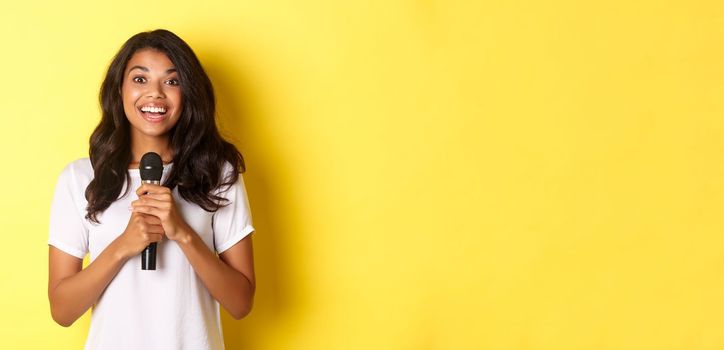 Portrait of happy african-american girl, looking amused while giving speech, holding microphone and smiling at camera, standing over yellow background.