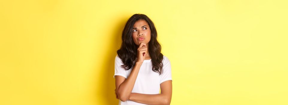 Portrait of attractive african-american woman thinking, making her choice, looking at upper left corner thoughtful and deciding, standing over yellow background.