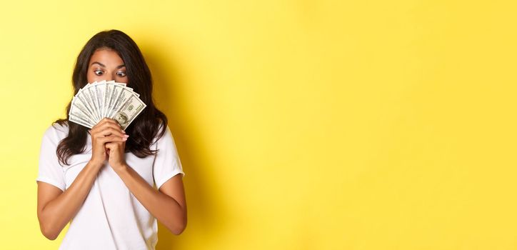 Portrait of lucky african-american girl winning money prize, holding cash and looking amazed, standing over yellow background.