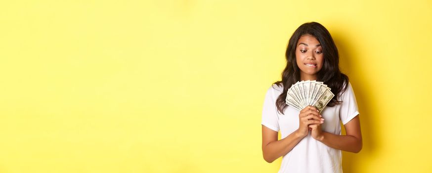 Portrait of attractive african-american woman, looking tempted at money, wanting to buy something, standing over yellow background.