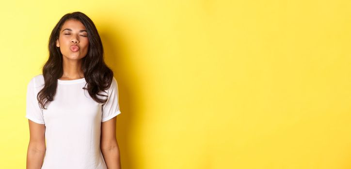 Image of beautiful and silly african-american girl, close eyes and waiting for kiss, standing in casual white t-shirt over yellow background.
