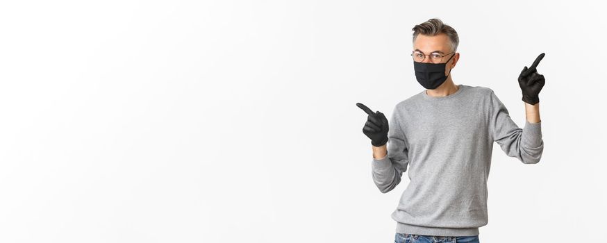 Concept of coronavirus, lifestyle and quarantine. Image of middle-aged man with grey hair, wearing black medical mask, gloves and glasses, pointing fingers sideways, showing copy space.