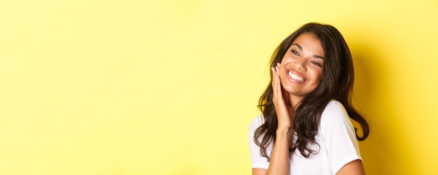 Image of gorgeous african-american woman touching her face, smiling pleased and looking left at copy space, standing over yellow background.