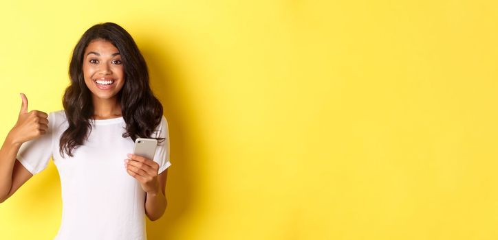 Portrait of young good-looking african american woman, showing thumbs-up while using mobile phone app, smiling pleased, standing over yellow background.