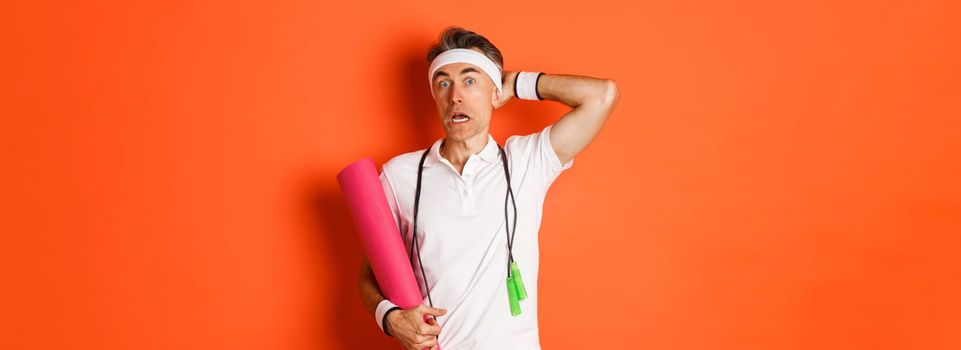 Concept of workout, gym and lifestyle. Image of awkward middle-aged athlete, looking confused, holding yoga mat and skipping rope, standing over orange background.