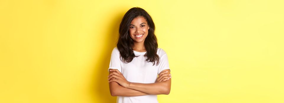 Image of happy smiling african-american girl in white t-shirt, cross arms on chest and looking confident, standing over yellow background.