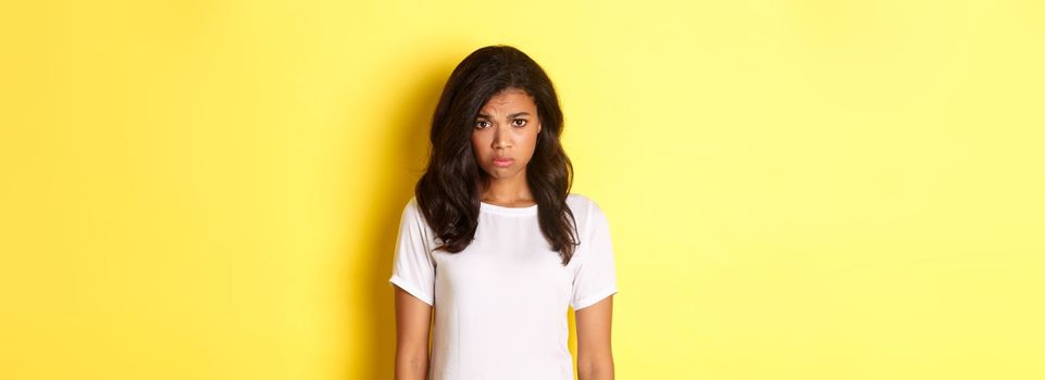 Portrait of gloomy and sad african-american girl, sulking and looking upset, standing in white t-shirt against yellow background.