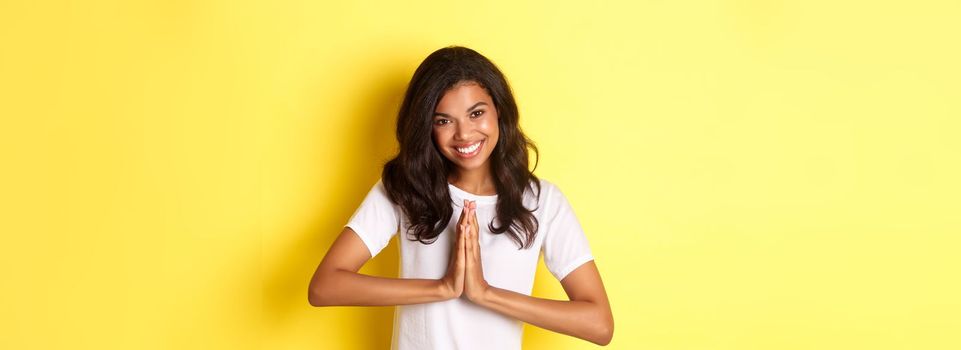 Image of cute african american woman, saying thank you and smiling, press hands together to express gratitude, standing over yellow background.