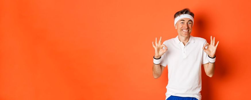 Concept of sport, fitness and lifestyle. Portrait of cheerful middle-aged male athlete, smiling pleased and showing okay signs, approve or recommend something, standing over orange background.