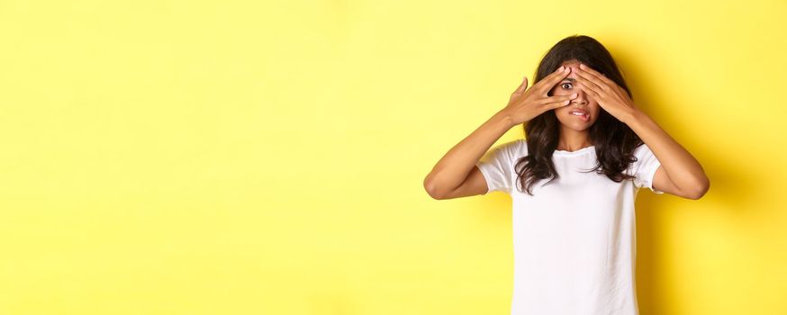 Portrait of embarrassed and shocked african-american girl, shut her eyes but peeking through fingers at something disturbing, standing over yellow background.