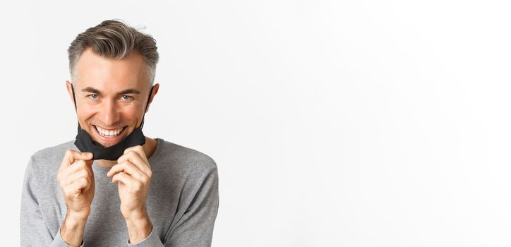 Close-up of handsome middle-aged man, taking-off medical mask and looking happy, smiling pleased, standing over white background. Concept of covid-19, social distancing and quarantine.