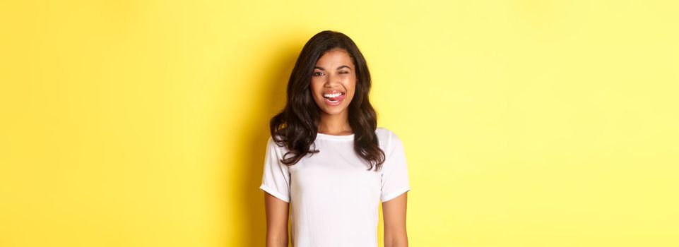 Image of carefree african-american girl in white t-shirt, showing tongue, smiling and winking happy, standing over yellow background.