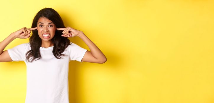 Portrait of annoyed and angry girl in white t-shirt, complaining on loud noise, shut ears and looking mad, standing over yellow background.