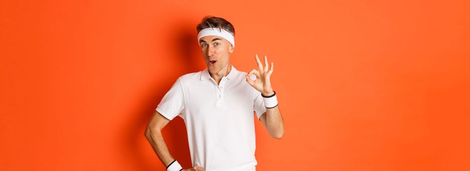 Concept of sport, fitness and lifestyle. Portrait of amazed adult man in workout uniform, showing okay sign and looking impressed, standing over orange background.