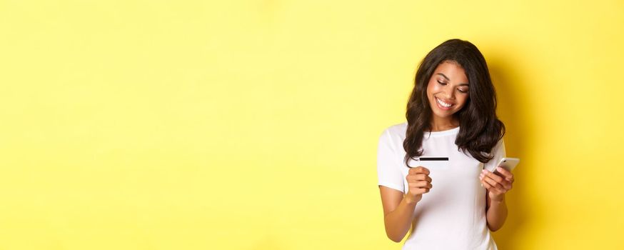 Young attractive woman shopping online, using mobile phone with credit card and smiling, standing over yellow background.