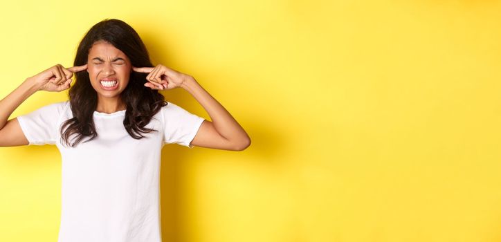 Portrait of annoyed and pissed-off african-american girl, shut her ears and grimacing from loud awful noise, being disturbed by something loud, standing over yellow background.