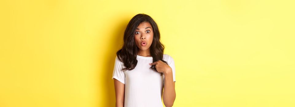 Portrait of surprised african-american girl, pointing finger at herself, standing over yellow background.