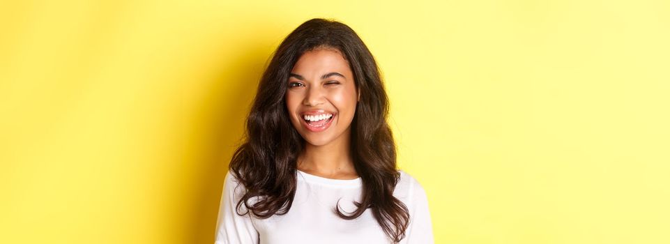 Close-up of cheerful african-american girl, winking and smiling at camera, recommending something good, standing over yellow background.