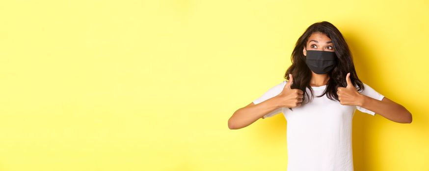 Concept of covid-19, social distancing and lifestyle. Image of cheerful african-american girl in black face mask, showing thumbs-up and looking happy at upper left corner, yellow background.