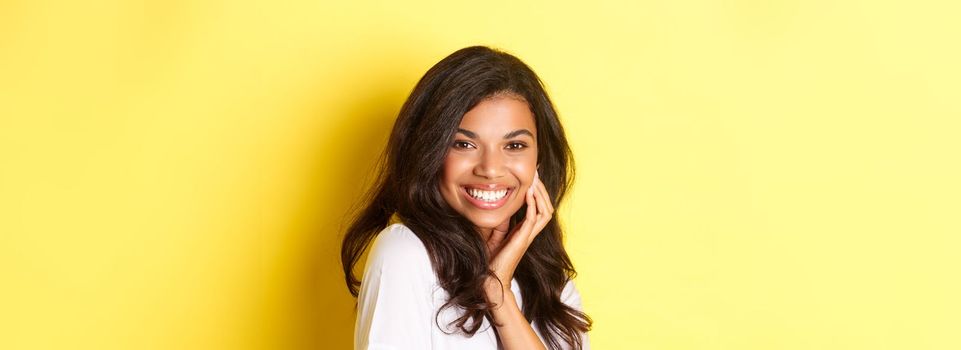 Image of beautiful african-american woman, smiling flirty and touching face, advertising beauty product, standing against yellow background.