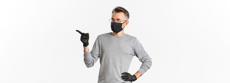 Concept of coronavirus, lifestyle and quarantine. Intrigued middle-aged man in medical mask, gloves and glasses pointing finger right and looking at advertisement, standing over white background.