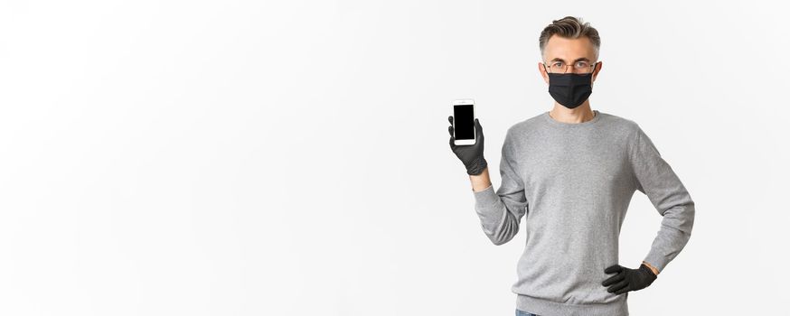 Concept of covid-19, social distancing and lifestyle. Image of confident middle-aged man in glasses, medical mask and gloves, showing smartphone screen, demonstrate an app.