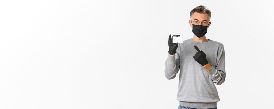 Concept of covid-19, social distancing and lifestyle. Handsome middle-aged man in medical mask, gloves and glasses, pointing finger at credit card, standing over white background.