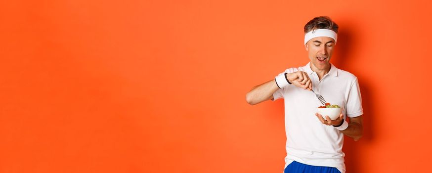 Concept of sport, fitness and lifestyle. Portrait of joyful middle-aged guy in workout uniform, holding fork and salad, eating healthy food, standing over orange background.