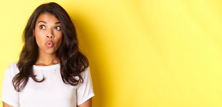 Close-up of impressed african-american woman in white t-shirt, saying wow, looking at upper left corner advertisement, standing over yellow background.