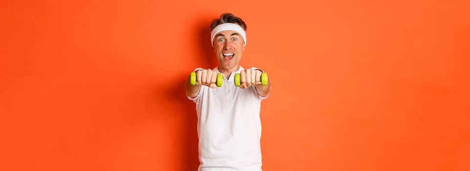 Image of active middle-aged fitness guy, doing sport exercises with dumbbells and smiling excited, standing over orange background.