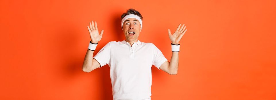 Portrait of surprised and happy middle-aged senior male exercises, raising hands up amazed and pleased, standing over orange background.