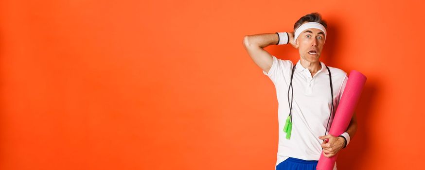 Concept of workout, gym and lifestyle. Image of awkward middle-aged athlete, looking confused, holding yoga mat and skipping rope, standing over orange background.