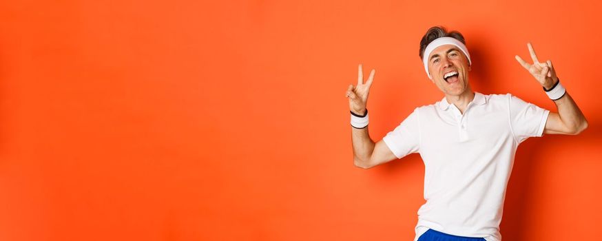 Image of happy and healthy middle-aged man in sport clothing, showing peace signs and smiling, workout over orange background.