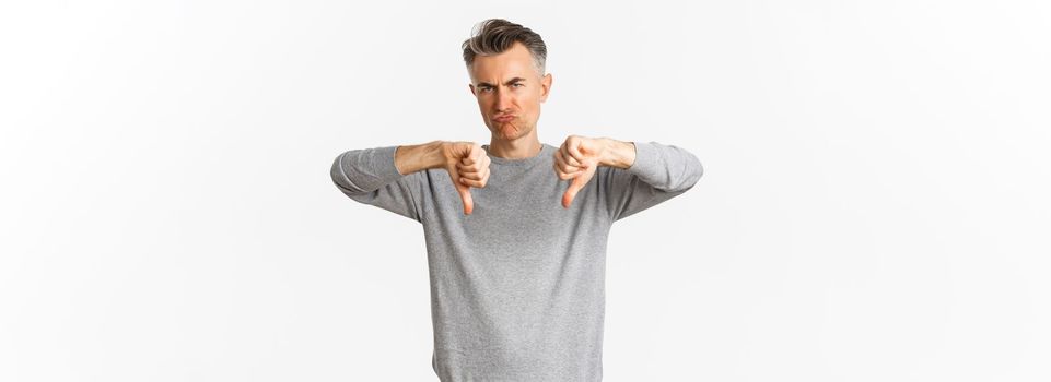 Skeptical and disappointed middle-aged man, grimacing unamused and showing thumbs-down, dislike something bad, standing over white background.