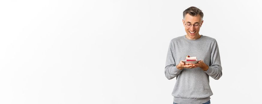 Portrait of handsome middle-aged man, looking happy at delicious cake, standing over white background.