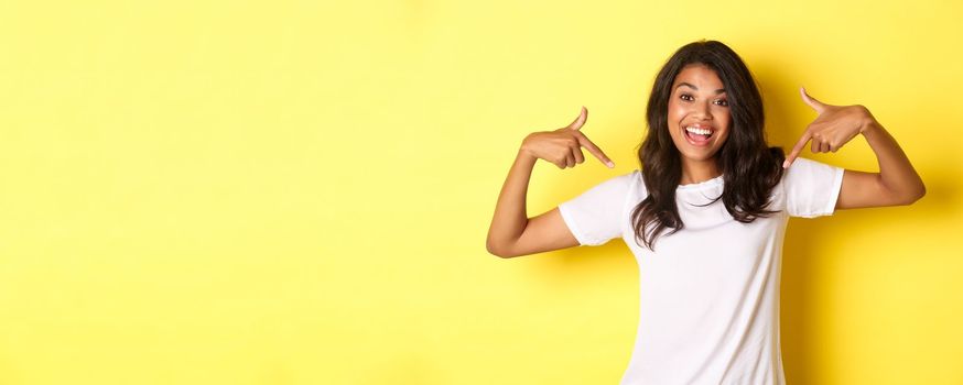 Portrait of cheerful african-american girl in white t-shirt, pointing fingers at your logo on center, standing over yellow background and smiling.