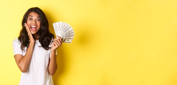 Image of excited lucky girl, winning money and smiling amazed, standing over yellow background.