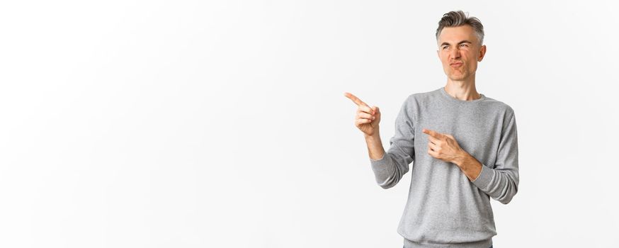 Portrait of skeptical handsome adult man, wearing grey casual sweater, grimacing as looking and pointing at something bad, express negative opinion, standing over white background.