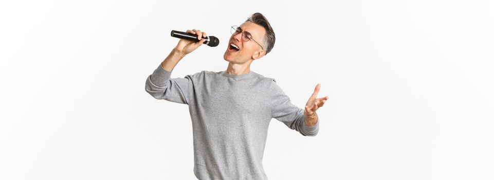 Portrait of passionate middle-aged man singing serenade in microphone, standing carefree over white background.