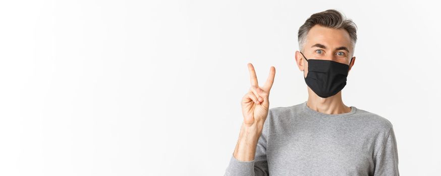 Concept of covid-19, social distancing and quarantine. Close-up of cheerful middle-aged guy in medical mask, showing peace sign and smiling, standing over white background.