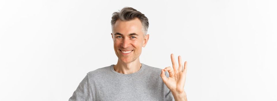 Close-up of handsome middle-aged man, showing okay sign and smiling, guarantee quality, assure that everything good, standing over white background.