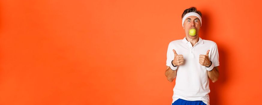 Image of funny middle-aged guy in fitness clothing, holding apple in mouth, showing thumbs-up, standing over orange background.