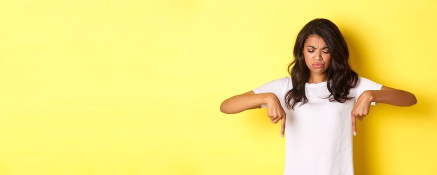 Portrait of sad and disappointed african-american girl, complaining about something bad, looking and pointing fingers down with displeased face, standing over yellow background.