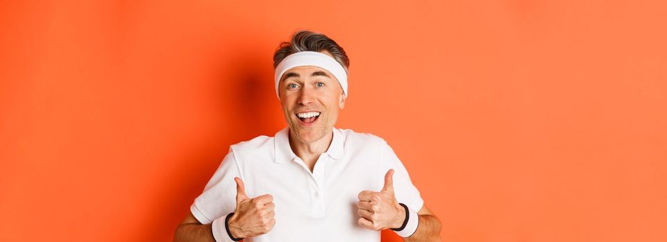 Close-up of cheerful middle-aged male athlete, wearing headband and t-shirt for workout, showing thumbs-up, approve something good, standing over orange background.