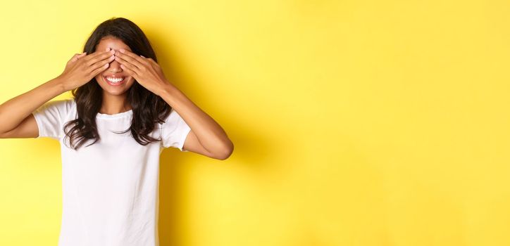 Image of excited african-american girl waiting for surprise, smiling and covering eyes with hands, standing over yellow background.