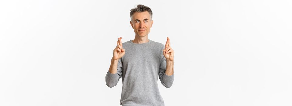 Image of hopeful middle-aged man making a wish, crossing fingers and looking upper left corner, standing over white background.