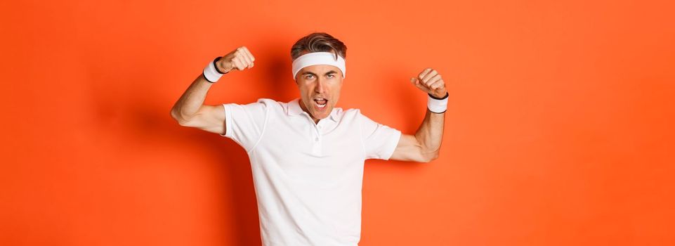 Image of funny middle-aged sportsman in white headband and t-shirt, flexing biceps to show-off, standing against orange background.
