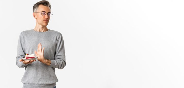 Portrait of displeased middle-aged man, looking away disgusted and rejecting piece of cake, standing bothered over white background.