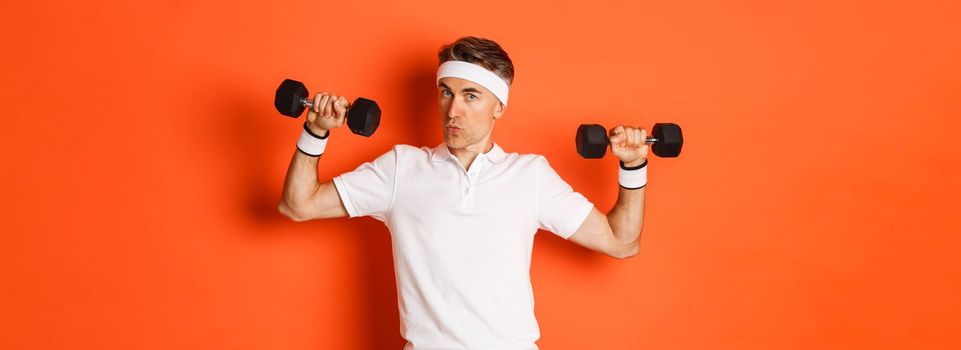 Concept of workout, gym and lifestyle. Image of healthy and strong middle-aged fitness guy, doing sport exercises with dumbbells, standing over orange background.
