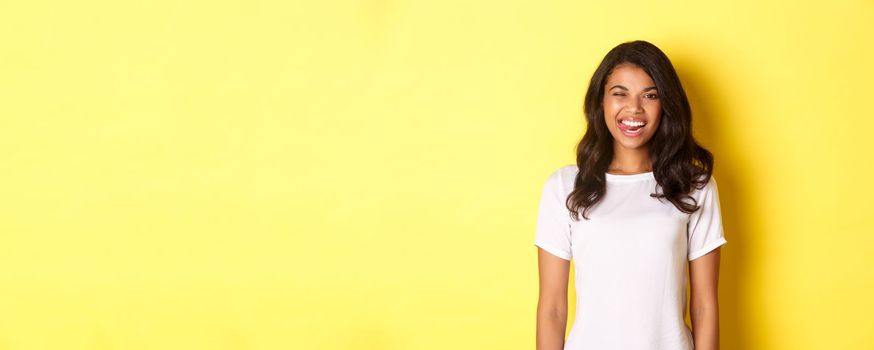 Image of carefree african-american girl in white t-shirt, showing tongue, smiling and winking happy, standing over yellow background.